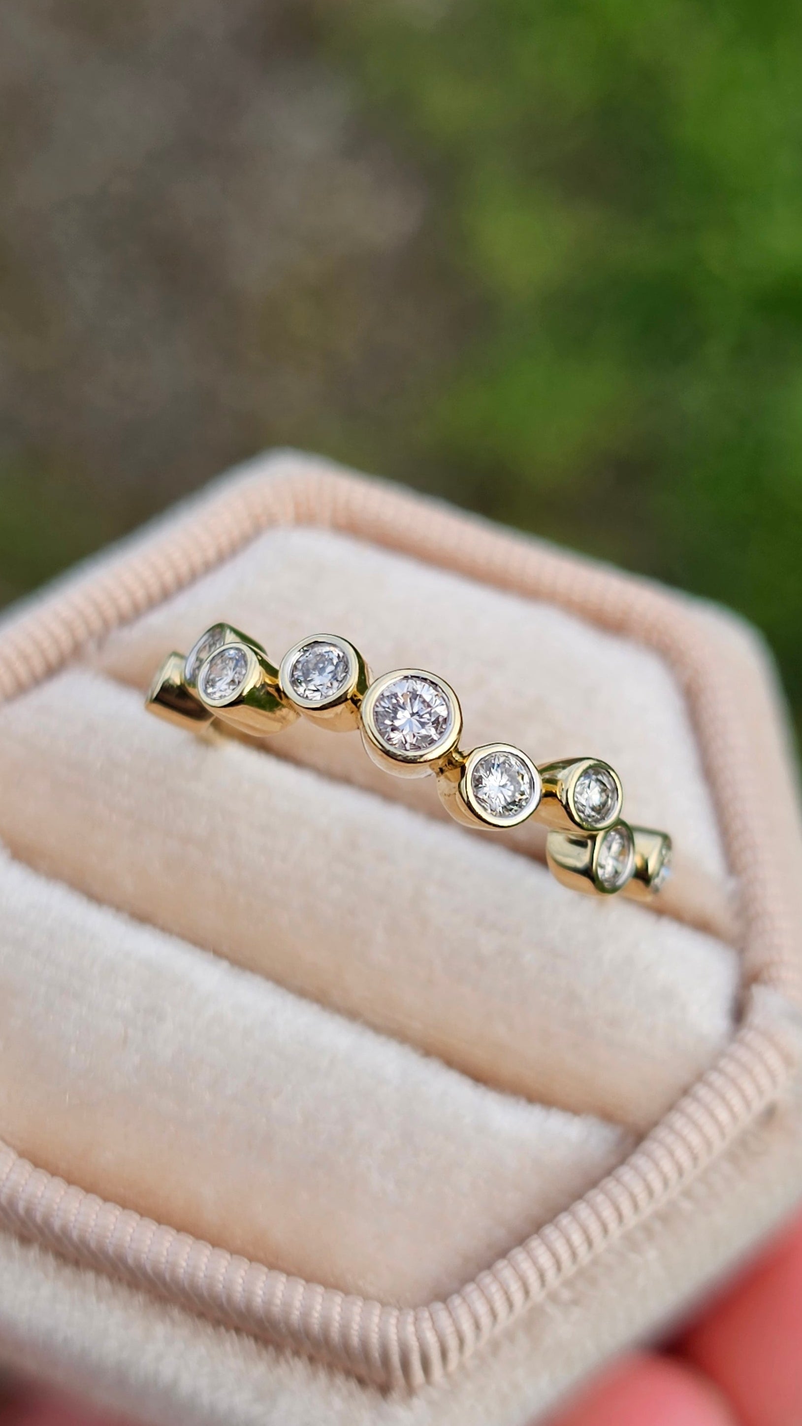 Gold ring with diamonds in a hexagonal beige box held by a hand against a blurred green background