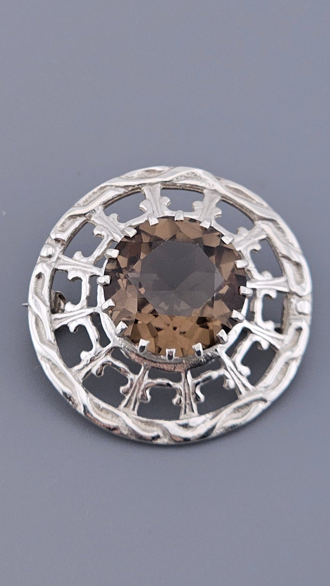 This exquisite vintage brooch is a beautiful example of traditional Scottish craftsmanship by the renowned silversmith Robert Allison. Centered with a large, faceted Smoky Quartz (often referred to in Scottish jewelry as a Cairngorm), the stone is secured in a multi-prong setting and framed by an intricate, openwork Celtic-inspired gallery.

Expertly crafted in 925 Sterling Silver, the brooch features a circular design with a textured border that captures the light beautifully. Fully hallmarked and restored